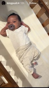 Newborn baby lying on a white bed, wearing a white onesie and gray pants, with a pacifier in its mouth.