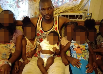 Man in a floral tank top sits on a sofa with several children, all with faces blurred, in a living room setting with floral curtains and an air conditioner behind them.