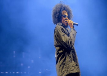 Male singer with an Afro hairstyle, wearing a dark coat and sunglasses, holding a microphone on a blue-lit stage.