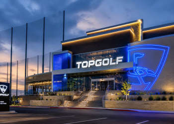 Exterior of a Topgolf venue at dusk, with a large illuminated blue sign and glass storefronts forming the entrance.