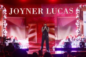 Male rapper in sunglasses performs on a red-lit stage with a JOYNER LUCAS backdrop above him.