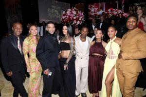 Celebrities posing for a group photo at a red-carpet event, with a large floral centerpiece and screens behind them reading 'Impact Awards' in gold lettering.