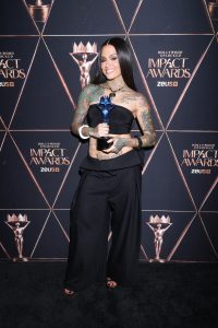 Woman with long dark hair and full-body tattoos smiles while holding a blue glass-trophy at the Impact Awards backdrop. Backdrop features geometric gold lines and 'Impact Awards' logos.