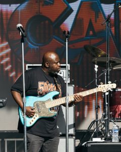 50YEAROFGOGO_shaunllewellyn__SHAUN LLEWELLYN_ROOTS PICNIC 26 DAY1_-13 - Baller Alert African American musician playing a blue bass guitar on a concert stage with microphones and a drum kit behind him