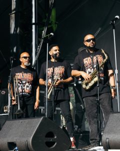 50YEAROFGOGO_shaunllewellyn__SHAUN LLEWELLYN_ROOTS PICNIC 26 DAY1_-18 - Baller Alert Three male musicians performing on an outdoor stage wearing matching black shirts that say '50 Years', with brass instruments (trombone and saxophone) and microphones in use.