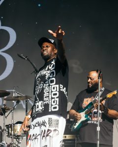 50YEAROFGOGO_shaunllewellyn__SHAUN LLEWELLYN_ROOTS PICNIC 26 DAY1_-23 - Baller Alert Man on stage pointing toward the audience while accompanied by a drummer and guitarist in a concert setting