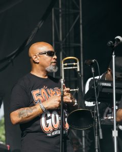 50YEAROFGOGO_shaunllewellyn__SHAUN LLEWELLYN_ROOTS PICNIC 26 DAY1_-9 - Baller Alert Musician wearing sunglasses on stage, holding a trombone beside a microphone.