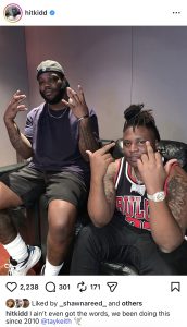 Two men sit on a black sofa making hand signs; one wears a dark T‑shirt and cap, the other wears a Chicago Bulls jersey with visible tattoos.
