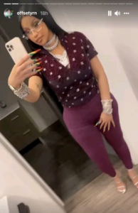 Person taking a mirror selfie wearing a burgundy patterned top, purple pants, rainbow nails, silver jewelry, clear glasses, and holding a white phone.