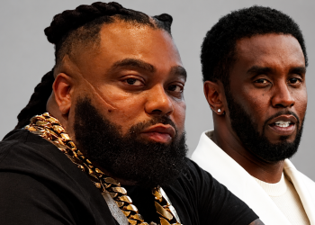 Two men pose together; left man wears heavy gold chains and has braided hair, right man wears a white blazer against a gray background.