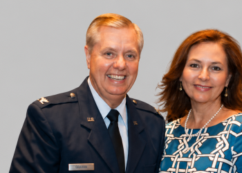 Lindsey Graham's sister Darline Graham Nordone with the senator in 2015 - Baller Alert Smiling man in a U.S. military dress uniform next to a woman in a blue patterned dress against a gray backdrop, both posing for a photo.