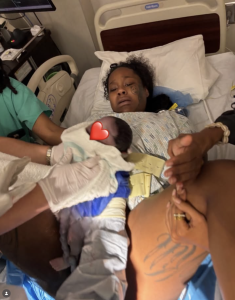 - Baller Alert Hospital scene: patient lying in bed holding a newborn baby wrapped in a patterned blanket, with a nurse nearby and medical equipment in view.