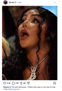 - Baller Alert Close-up of a surprised woman with long dark curls, wearing a diamond necklace with a crown-shaped 'B' pendant.