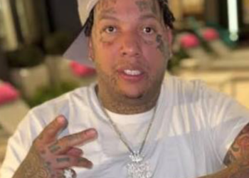 Close-up of a man with facial tattoos wearing a white shirt, making a peace sign toward the camera.