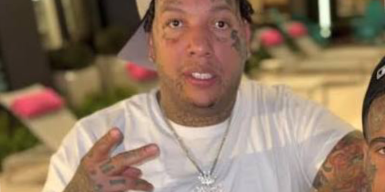 Close-up of a man with facial tattoos wearing a white shirt, making a peace sign toward the camera.