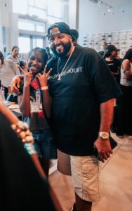 Smiling man in a black T-shirt and backwards cap posing with a girl who is flashing peace signs at an indoor event.