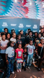 Group of children and young adults posing for a photo at a charity event in front of a blue backdrop with 'We The Best Foundation' and 'Snipes' logos.