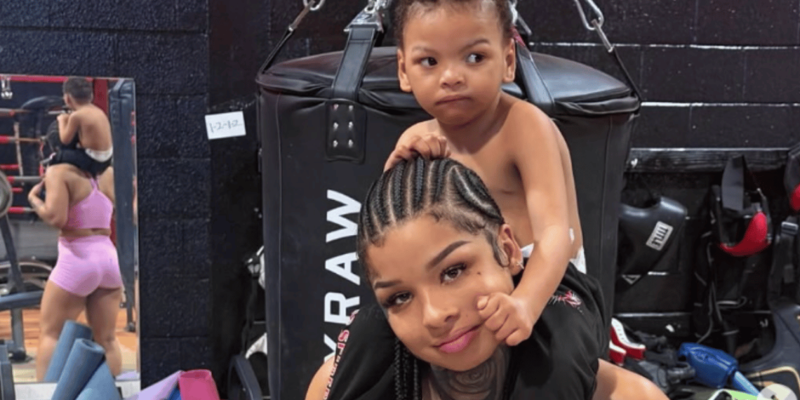 Woman with cornrow braids carries a toddler on her shoulders in a boxing gym next to a large punching bag, both looking to the side.
