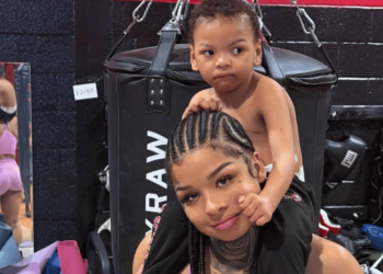Woman with cornrow braids carries a toddler on her shoulders in a boxing gym next to a large punching bag, both looking to the side.