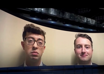 Two men in suits appear on a curved screen backdrop, as if on a video screen in a studio setting.