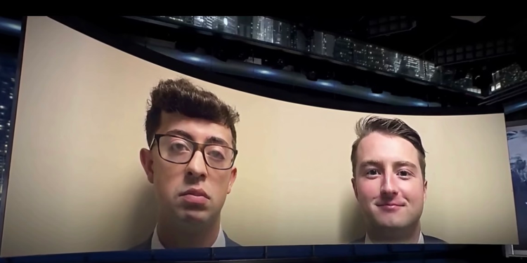 Two men in suits appear on a curved screen backdrop, as if on a video screen in a studio setting.