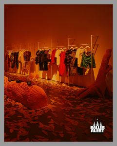 Clothes hung on racks in an orange-lit gallery, with leaf-covered floor and a large tree-root sculpture on the right.
