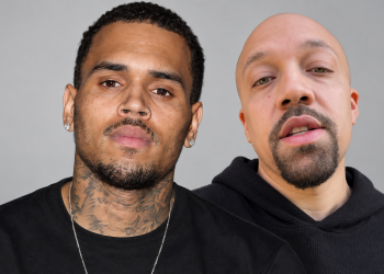 Two men posing for a close-up portrait: on the left, a tattooed man with a nose ring and earrings; on the right, a bald man with a goatee wearing a black hoodie.