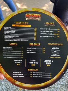 Round black menu board with a yellow-orange rim from a street eatery called No Knife Chicken & Waffles; lists waffles, mains, sides, egg rolls, signature sauces, fried fish & grits, crab fried rice, lobster & grits, and beverages with prices.