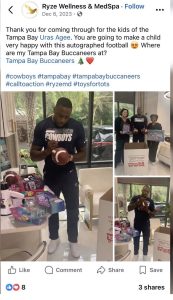 Man in a Dallas Cowboys shirt signs a football at a toy-donation setup, with toys and a Toys for Tots box nearby in a bright room.