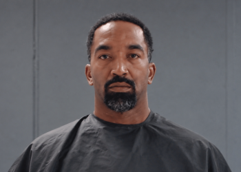 Portrait of a man with short curly hair and goatee wearing a black barber cape in front of gray studio panels, looking straight ahead.