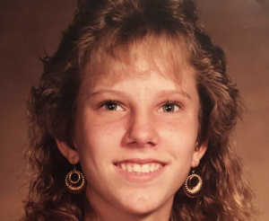 Smiling young woman with curly hair wearing gold circular earrings, facing the camera in warm lighting.