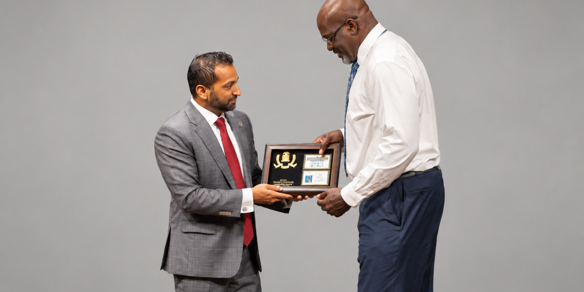 Two men in business attire exchange a framed award plaque against a gray backdrop.