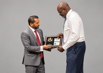 Two men in business attire exchange a framed award plaque against a gray backdrop.