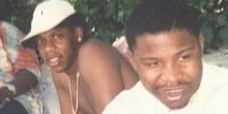 Two friends posing for a photo outdoors; one in a white shirt smiles at the right, with another person wearing a light cap in the background.