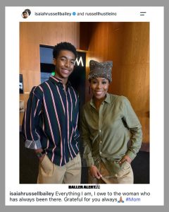 Two smiling people posing indoors in front of wood panels; man wears a striped shirt and watch, woman wears an olive shirt and a textured hat.