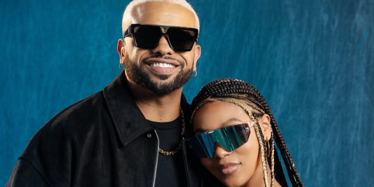 Two people pose together: a man with blond hair wearing large black sunglasses and a black jacket, smiling beside a woman in a mirrored, futuristic visor against a blue backdrop.