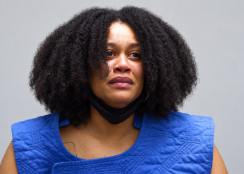 Portrait of a woman with curly hair, blue quilted vest, and a black mask pulled under her chin, looking slightly distressed against a gray backdrop.