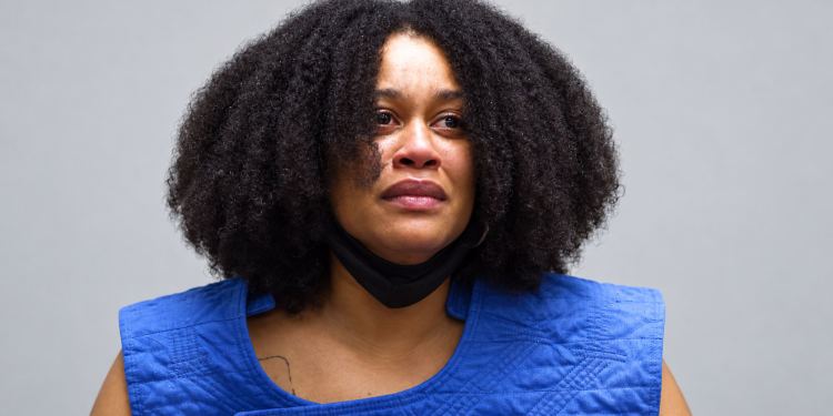 Portrait of a woman with curly hair, blue quilted vest, and a black mask pulled under her chin, looking slightly distressed against a gray backdrop.