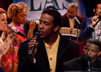 Five singers on a stage performing with microphones; central man in a yellow shirt and black blazer sings.