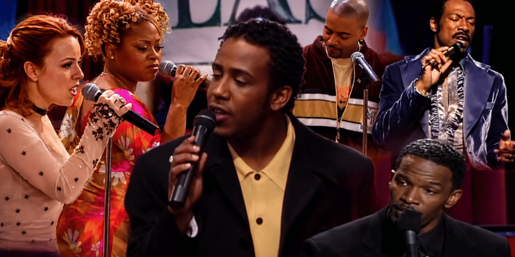 Five singers on a stage performing with microphones; central man in a yellow shirt and black blazer sings.