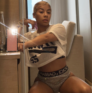 Person posing in a mirror selfie, wearing a white mesh top and glittery shorts with a Dolce & Gabbana waistband, seated beside a lamp.