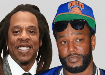 Two men posing together; left man with dreadlocks and a suit smiles, right man wearing a blue Knicks cap, sunglasses, and a beard.