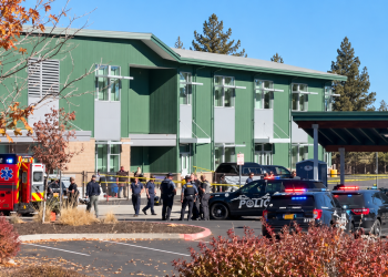 Emergency scene outside a green multi-story building with police cars and an ambulance; officers and bystanders behind caution tape.