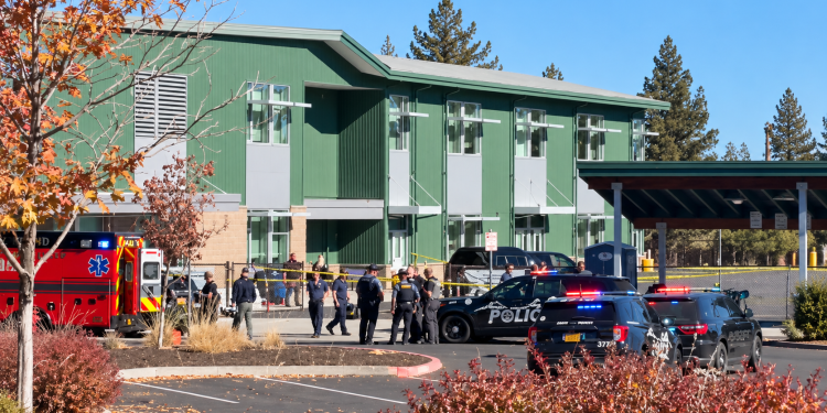 Emergency scene outside a green multi-story building with police cars and an ambulance; officers and bystanders behind caution tape.